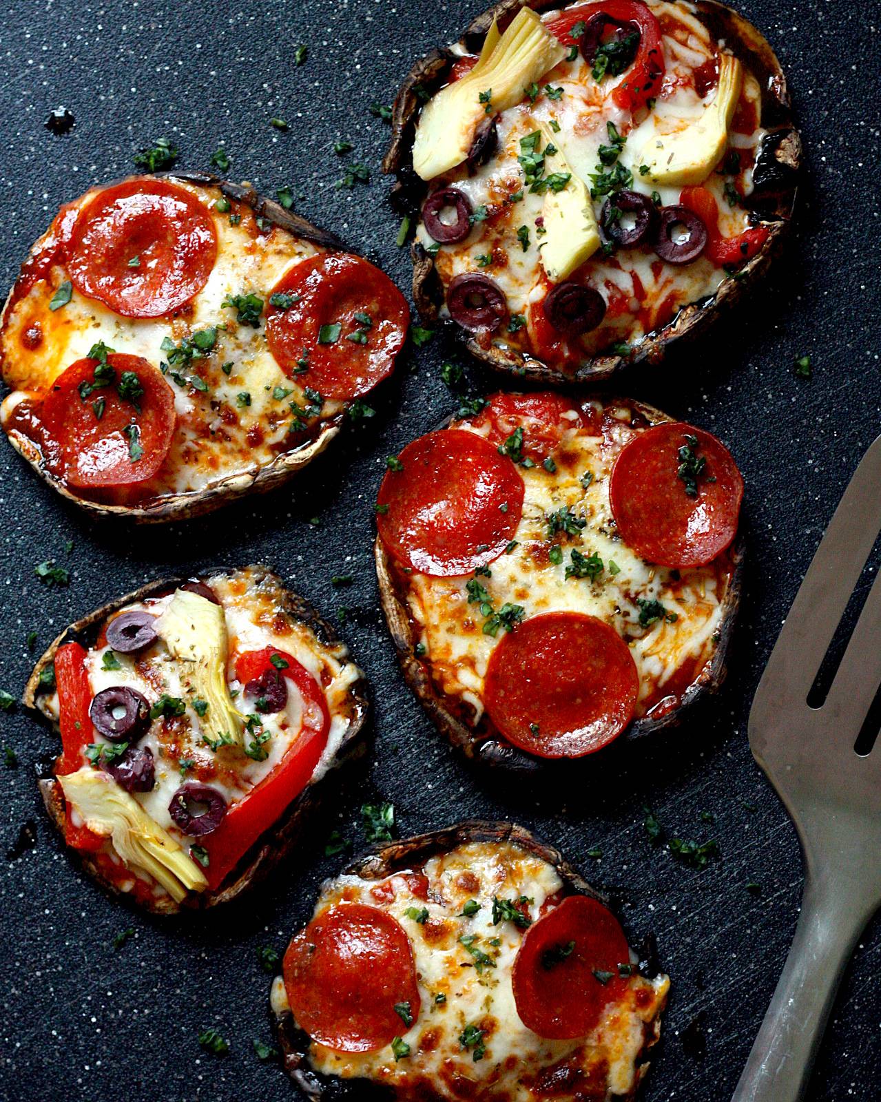 A black cutting board topped with portobello mushrooms which have been made into pizzas and topped with sauce, cheese and pepperoni.